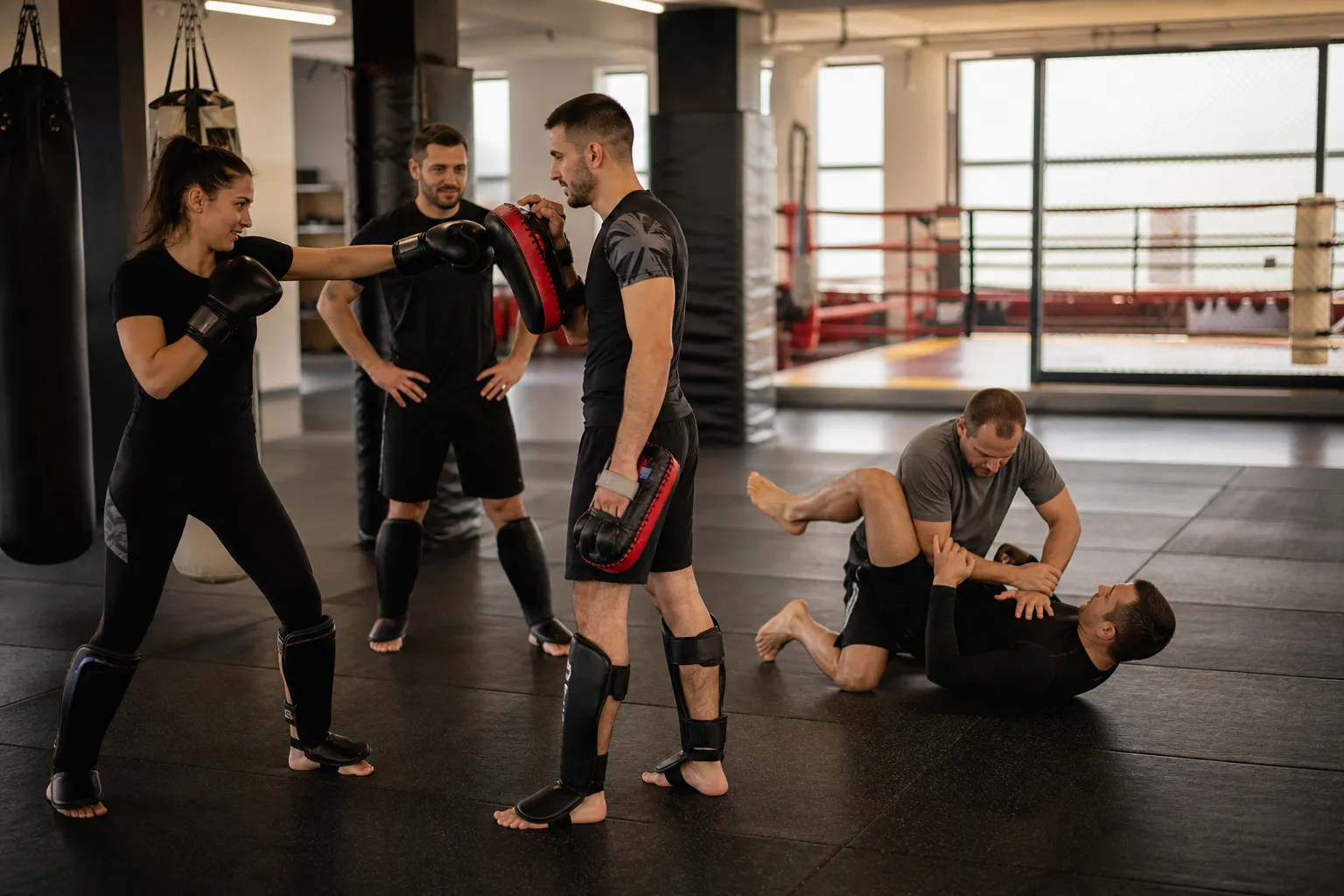 Adulți începători exersează controlat kickboxing și grappling pe saltele, într-o sală de arte marțiale.