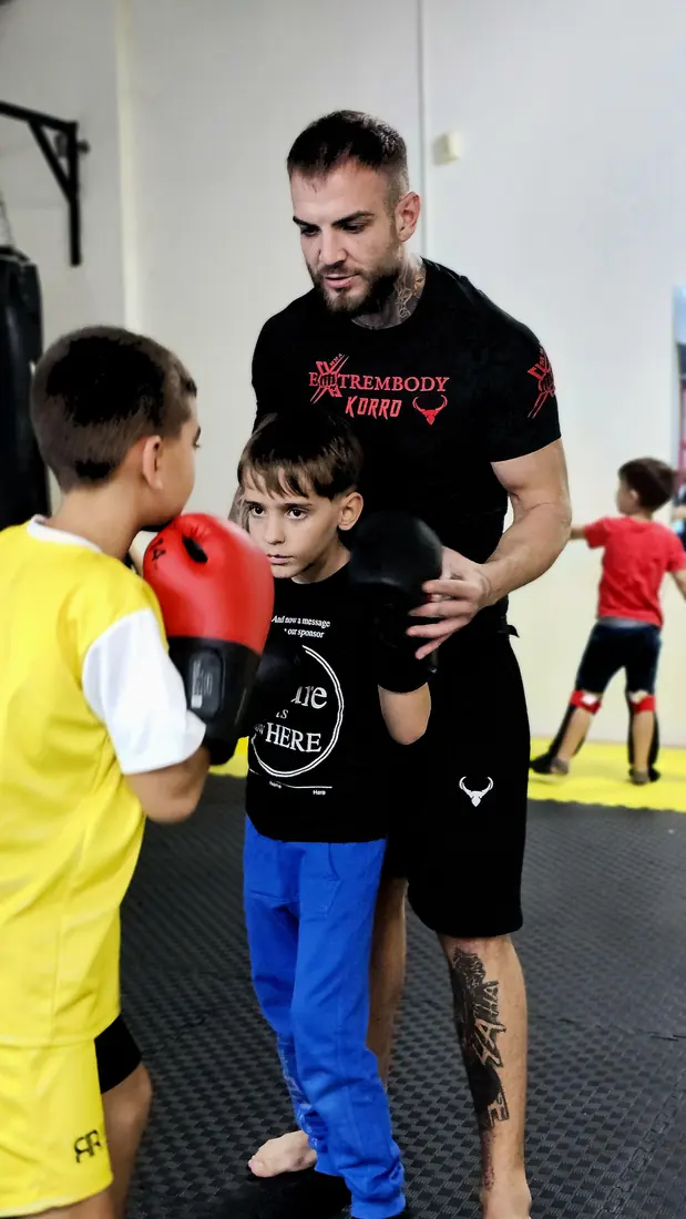Copil care exersează o lovitură într-o pernă ținută de antrenor, la un antrenament de kickboxing